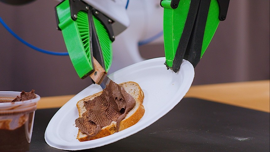 Robot using a kitchen tool to spread chocolate on a piece of bread, while holding the plate